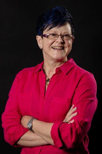 A woman with blue cropped hair and red shirt smiles at the camera with her arms folded