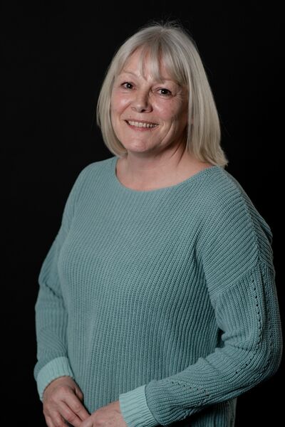 A woman in a teal jumper with a grey bob smiles warmly at the camera