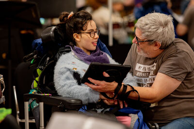 A young woman sitting in a wheelchair wearing a blue fluffy cardigan is smiling at her carer showing her a tablet