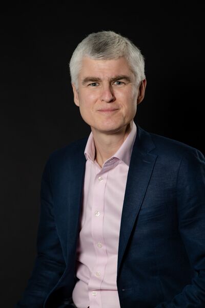 A man in a navy suit jacket and pale pink shirt with grey hair smiles at the camera
