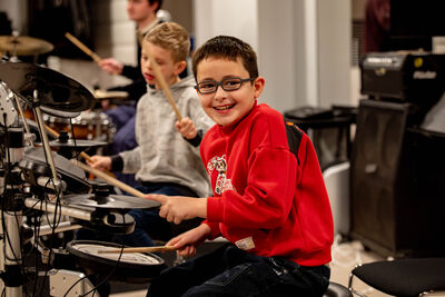A boy in red sweatshirt smiles whilst playing drums